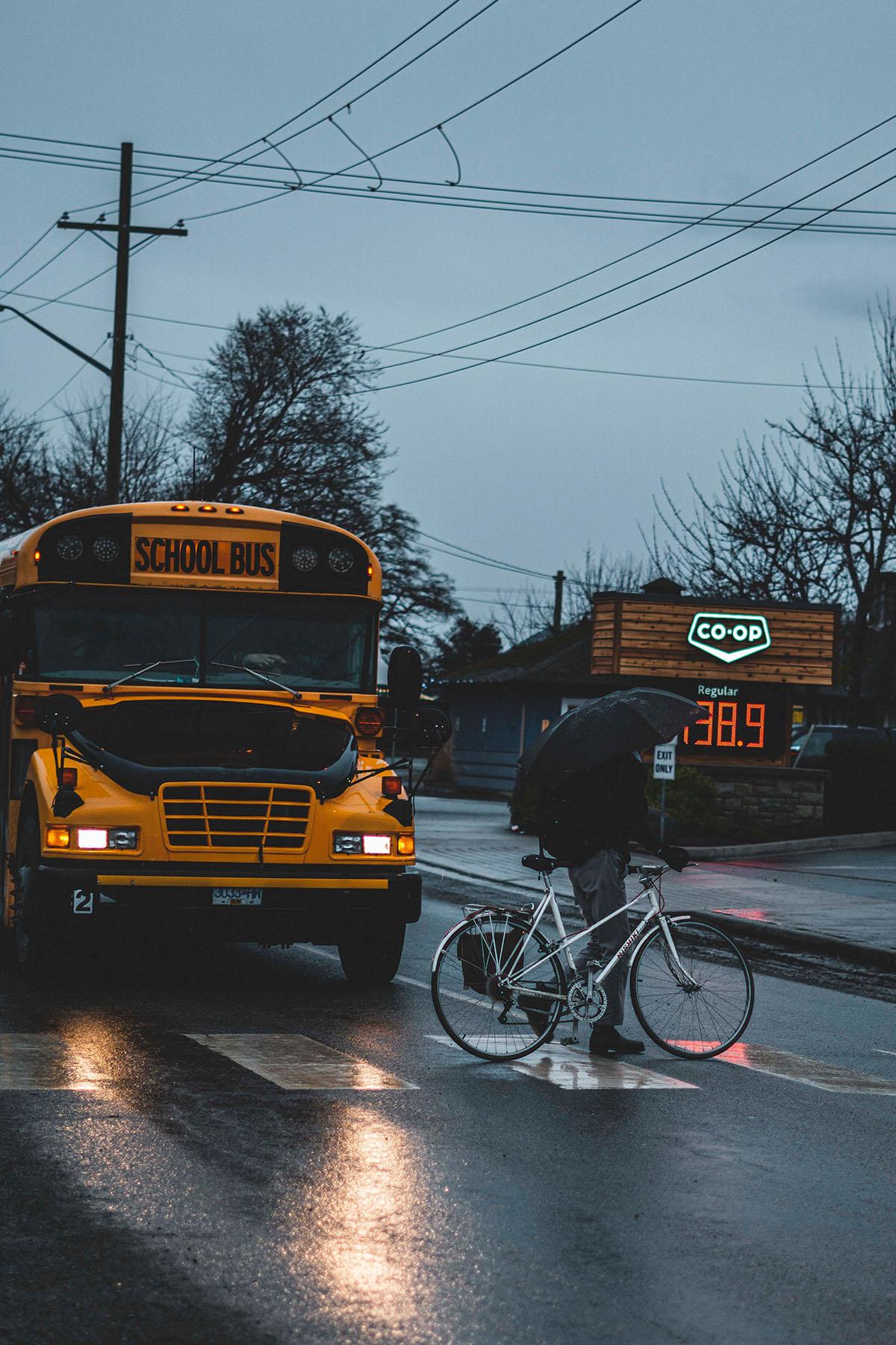 School bus stopped and a person on the crosswalk with a bike and umbrell.